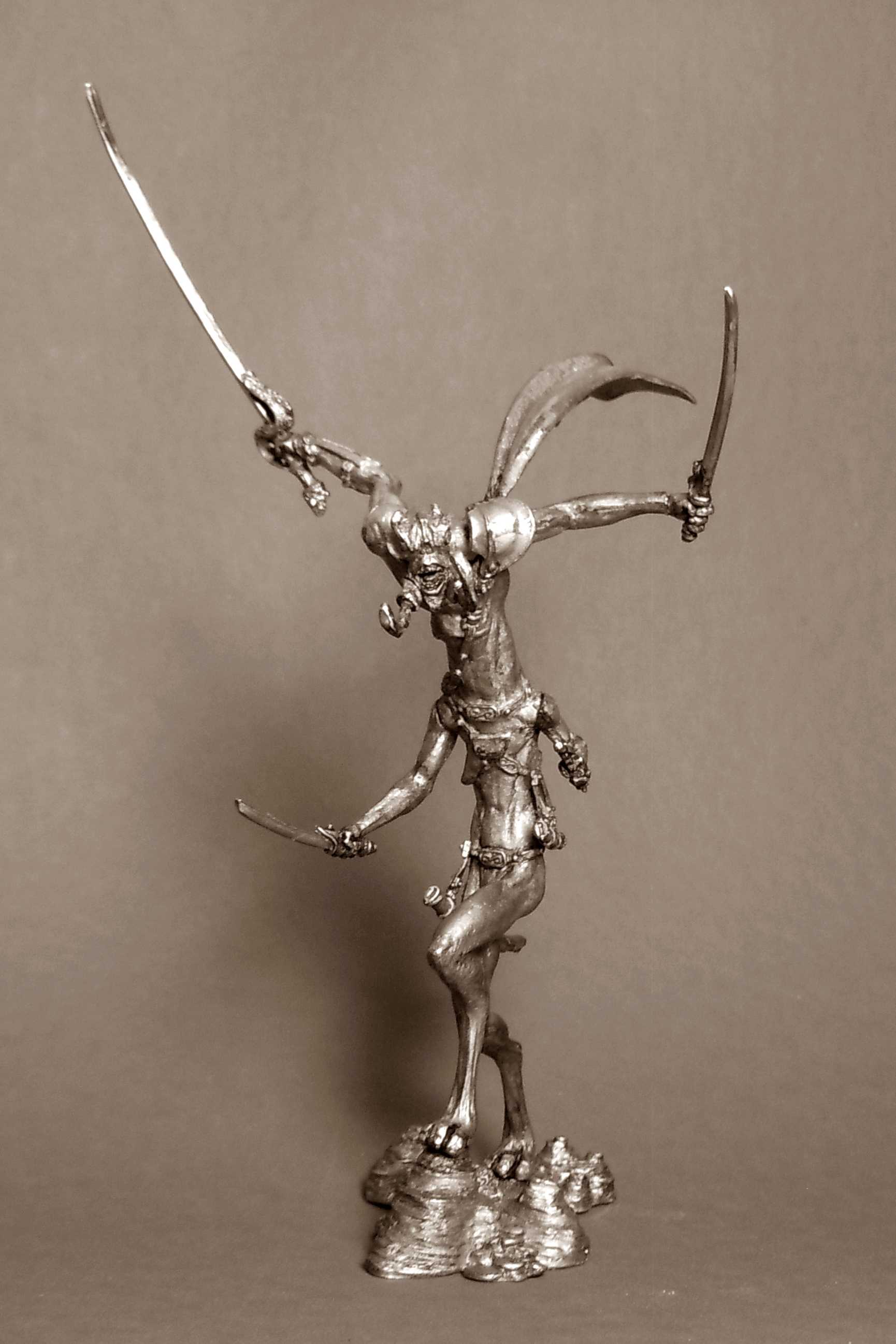 Unpainted metal miniature figure of a fantastical winged, horned creature wielding multiple curved swords, standing on a plain surface with dramatic side lighting.