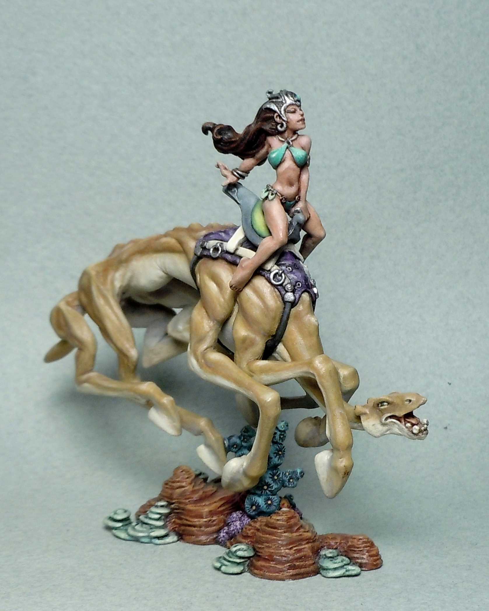 A painted miniature of a woman in a bikini-style outfit riding a leaping dog-like creature on a rocky base, with warm earthy tones.